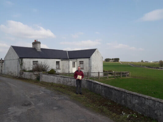 a man stands outside a house with a book in his hand County Mayo author