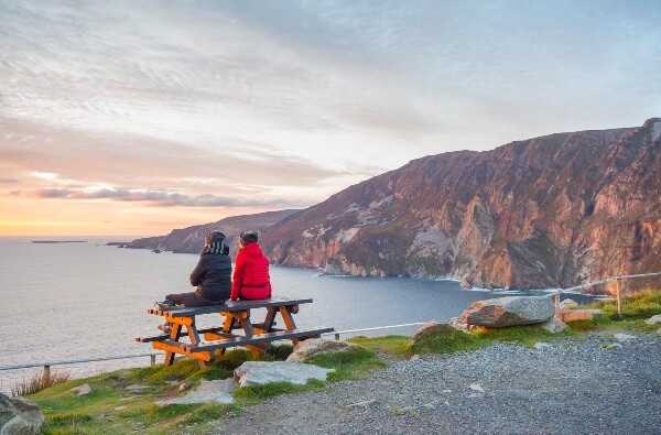 two people sitting on a bench looking at the ocean 10 undiscovered whiskey distilleries in Ireland