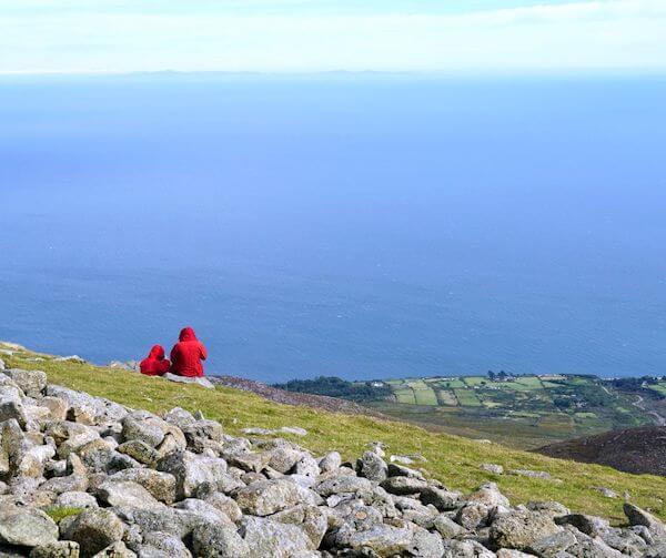 two people sitting on a mountain top looking at the ocean from Belfast to the Mourne Mountains